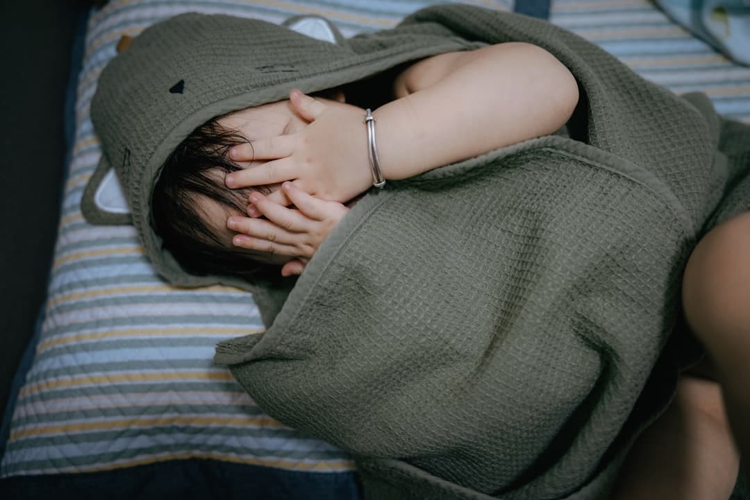 Child hides face under hooded towel on striped bed